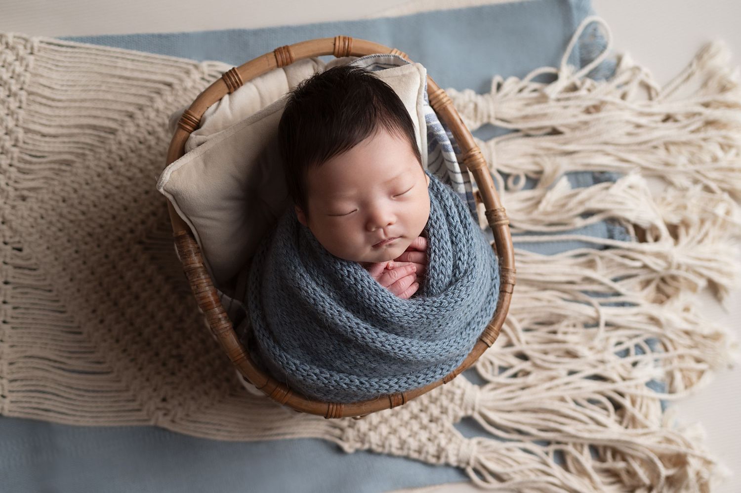 Sleeping baby nestled in blue knit wrap in rustic basket with fringed blanket.