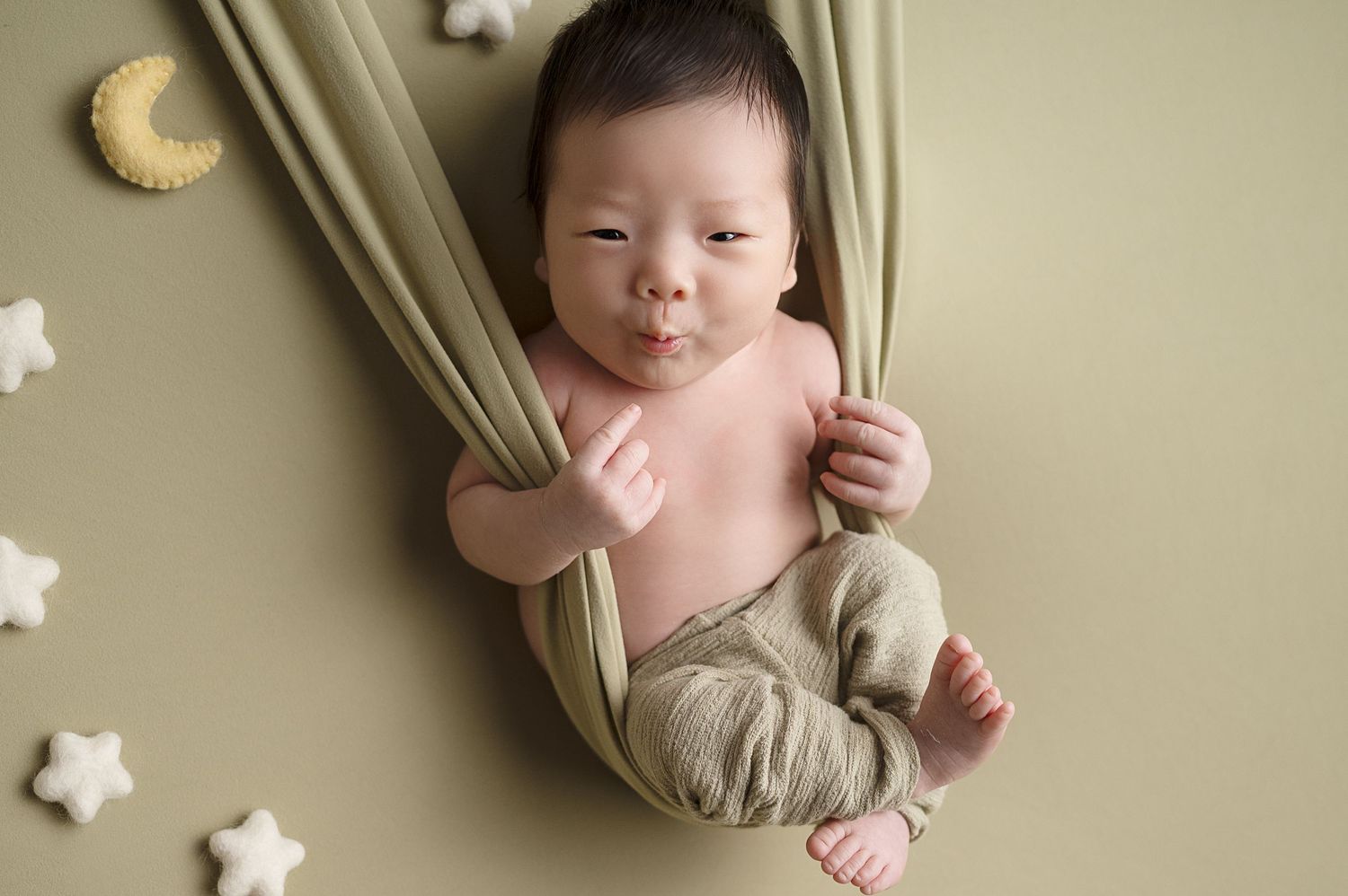 Newborn sleeping peacefully in olive fabric hammock with star and moon decorations.