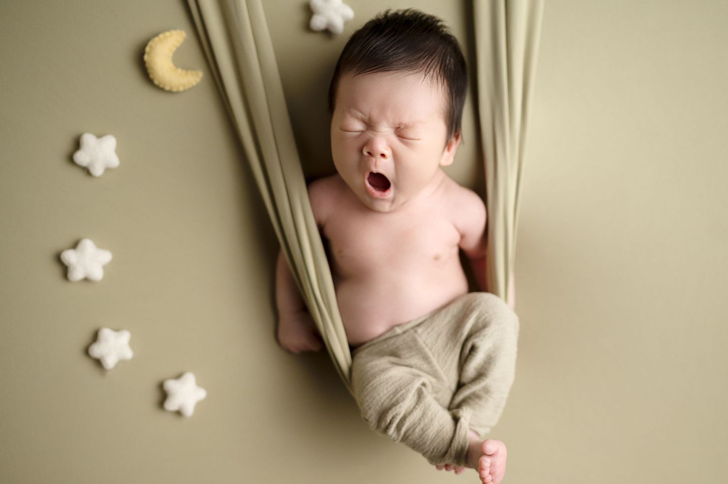 Yawning newborn lounging in sage green fabric with white star decorations on wall.