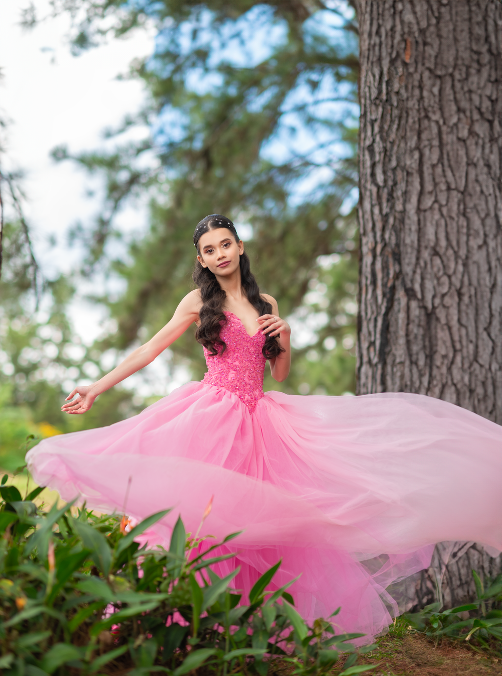 Pink ball gown swirls dramatically in a forest photoshoot with pine trees and flowers.