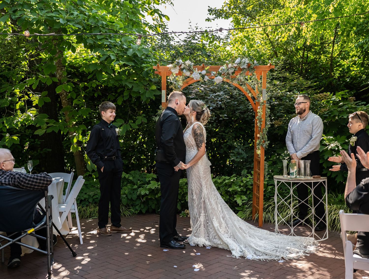 An intimate outdoor garden wedding ceremony under a wooden arch adorned with flowers and greenery.