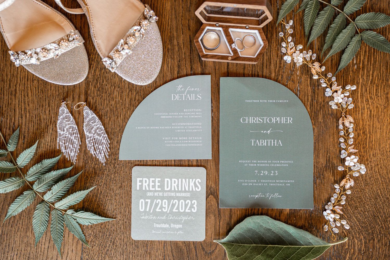 Wedding stationery set featuring sage green invitations and pearl sandals on wooden surface.