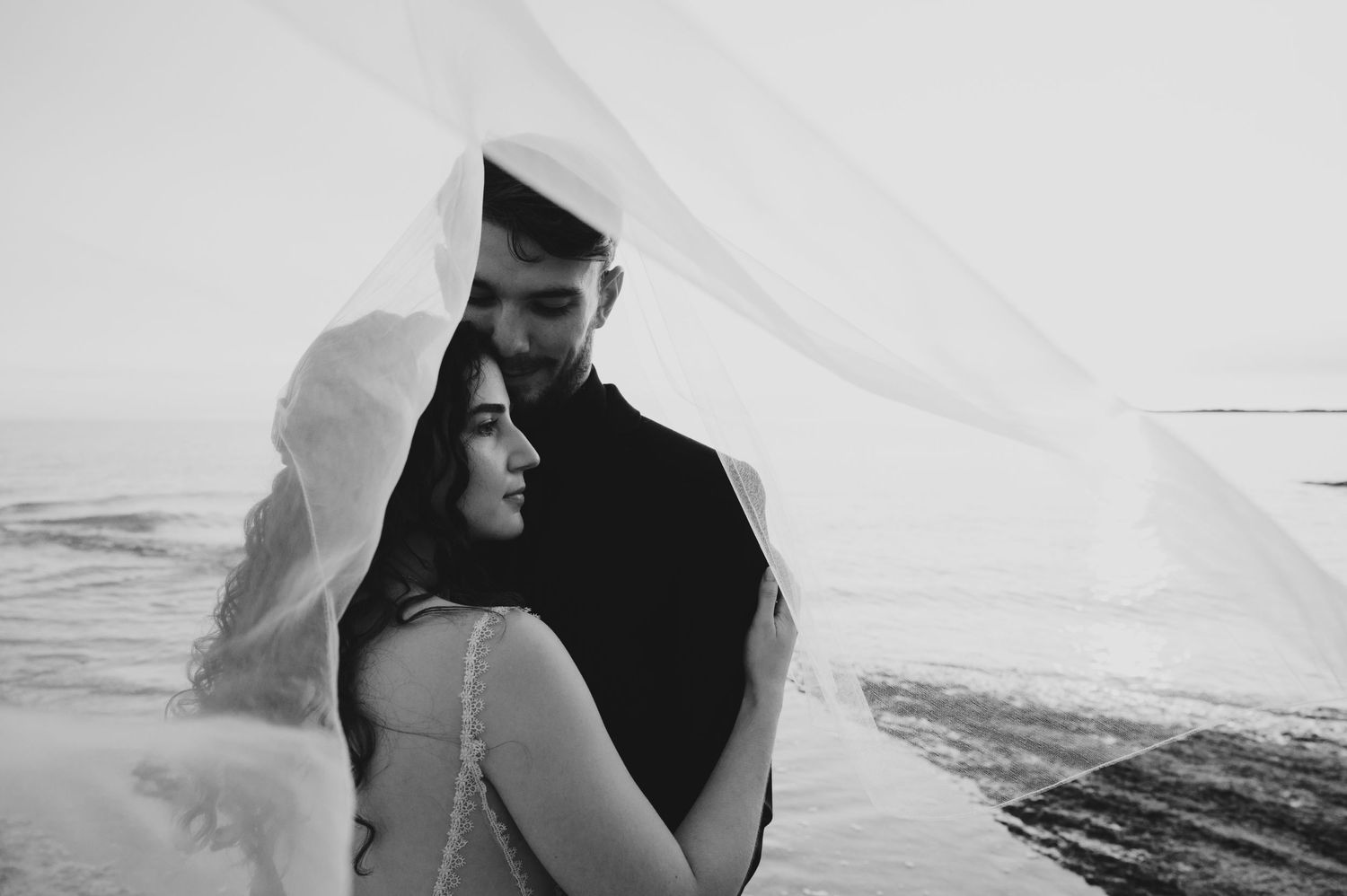 Black and white photo of bride and groom under flowing veil by the ocean.