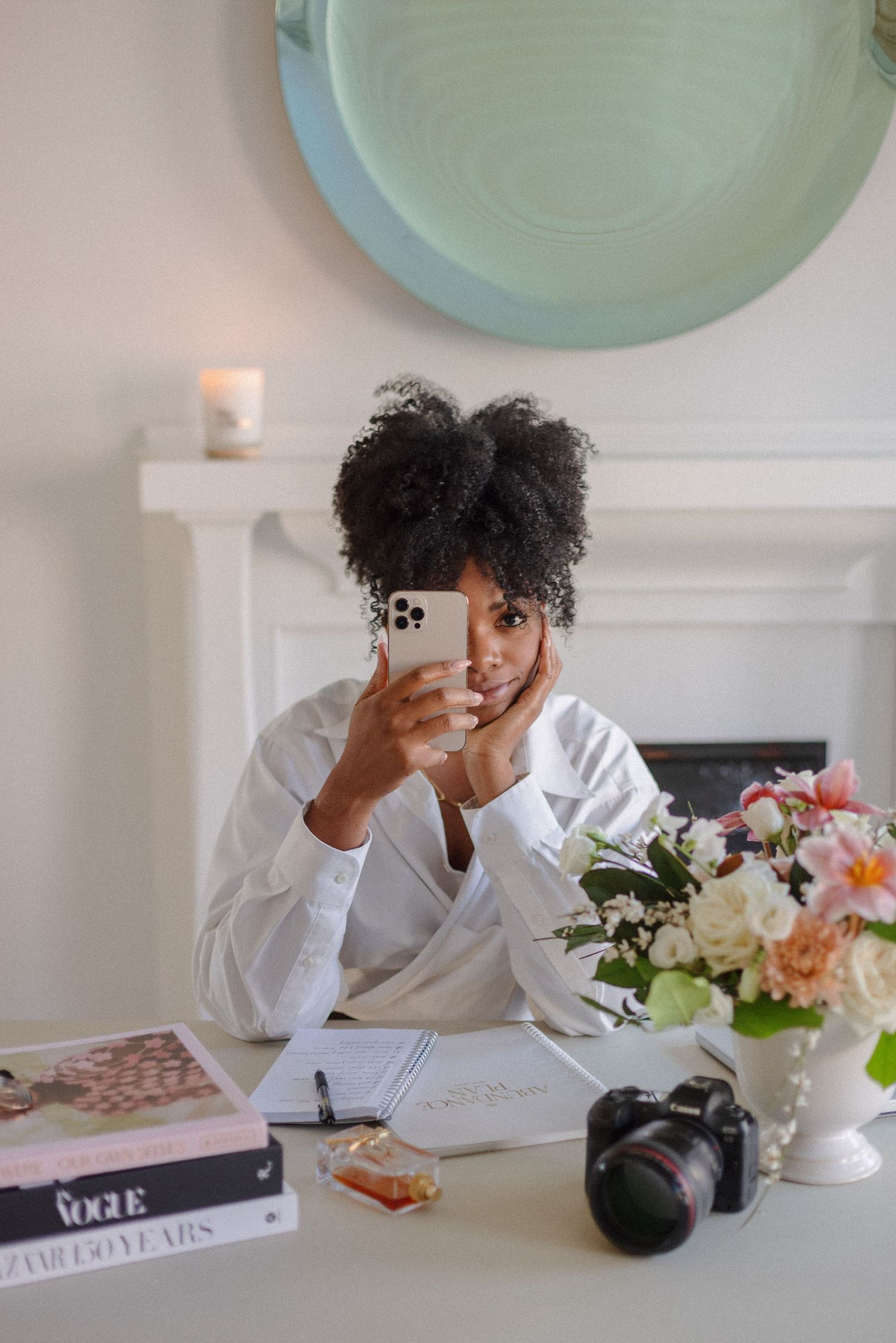 Content creator taking selfie at desk with flowers and camera equipment.