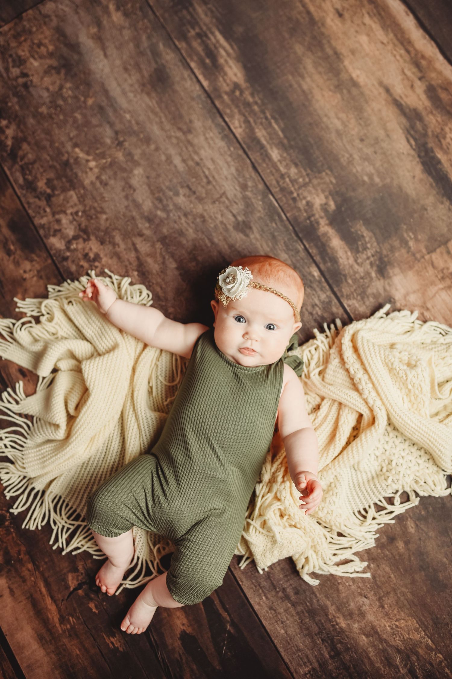 Infant wearing sage green romper lying on cream knit blanket on wooden floor.