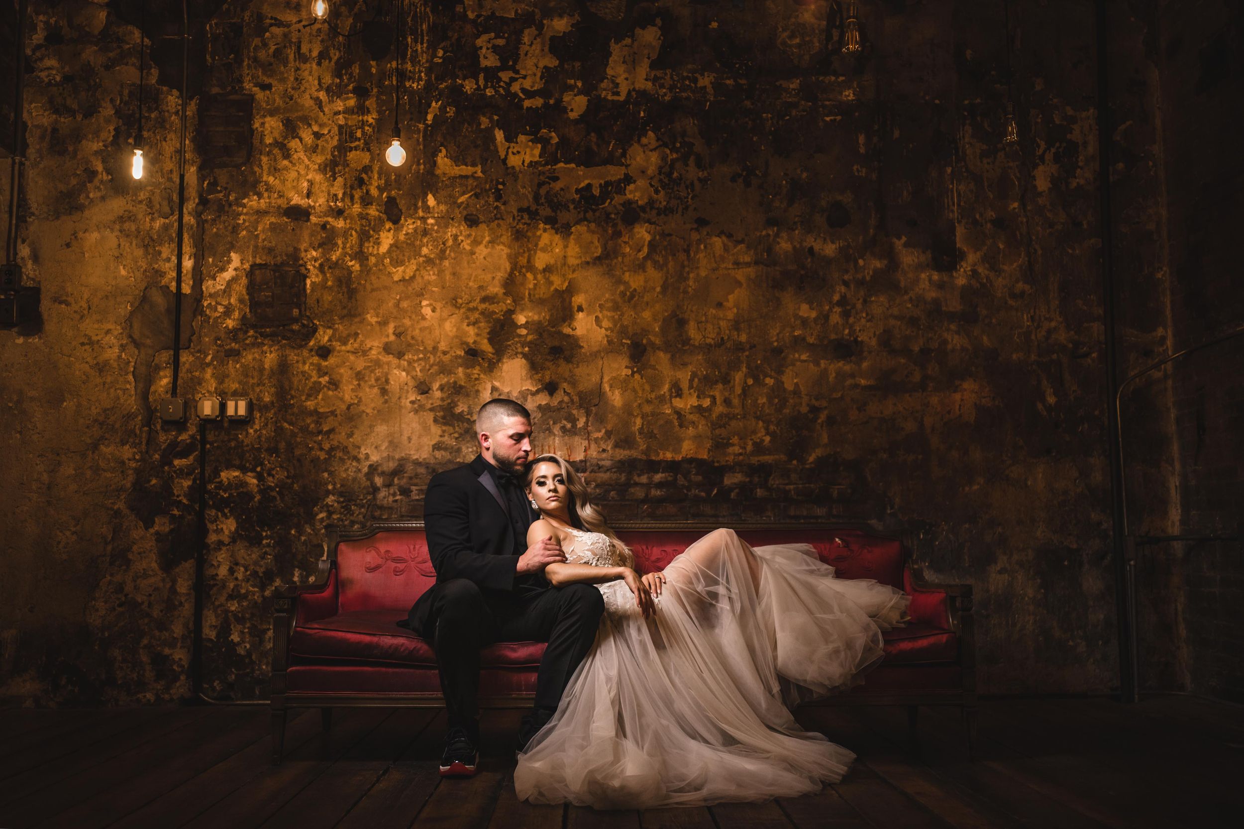 A romantic bride and groom rests on a vintage red sofa in a rustic industrial setting with warm lighting and textured walls
