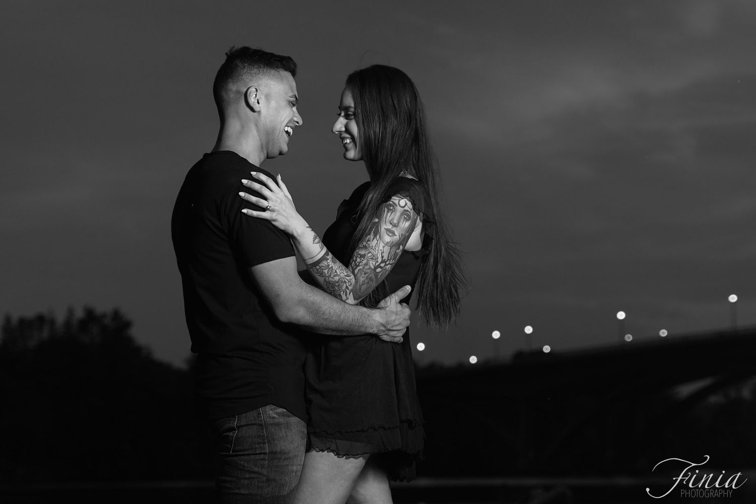 A romantic black and white portrait of a couple embracing at twilight with city lights glowing in the background.