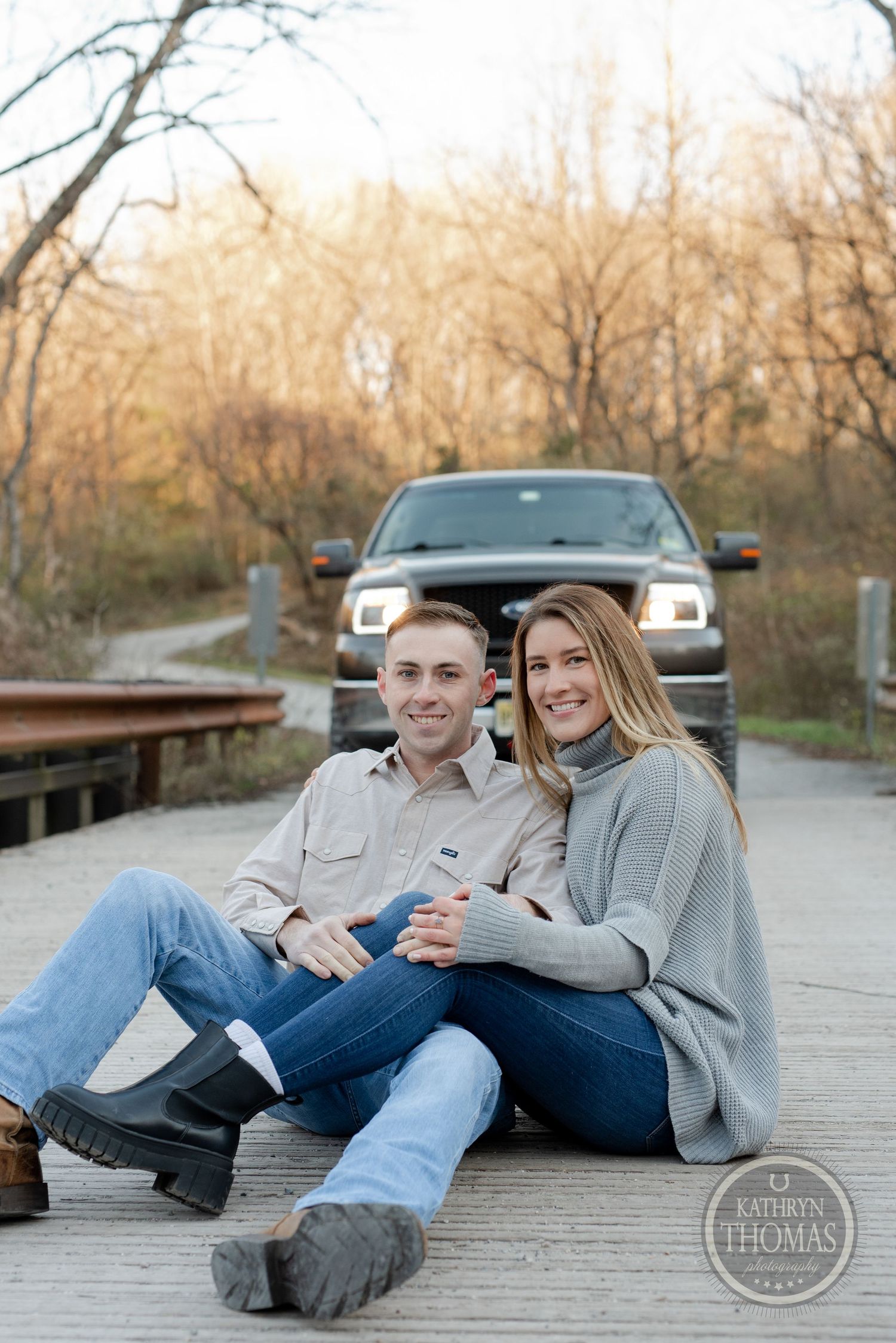 Casual couple sitting together on a wooden bridge with their truck in the background at sunset.