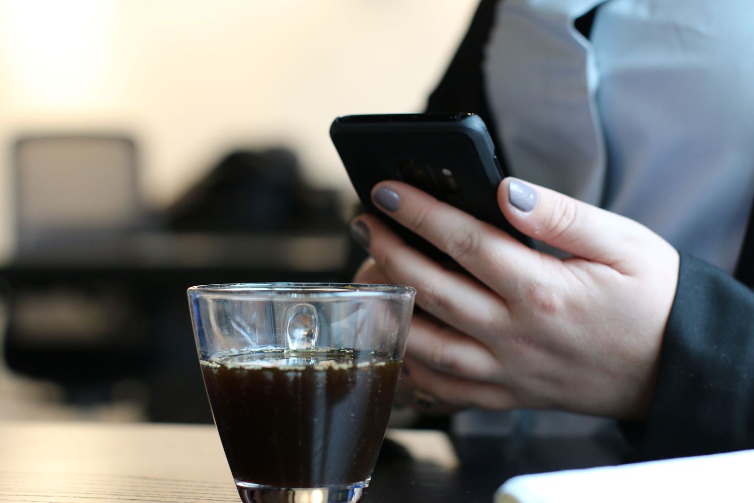 Hands holding smartphone next to glass of black coffee on wooden table.