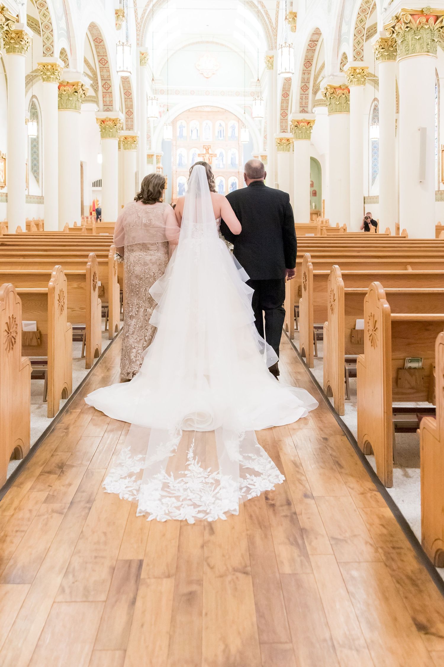 Bride in flowing white gown walks down church aisle with veil trailing behind.