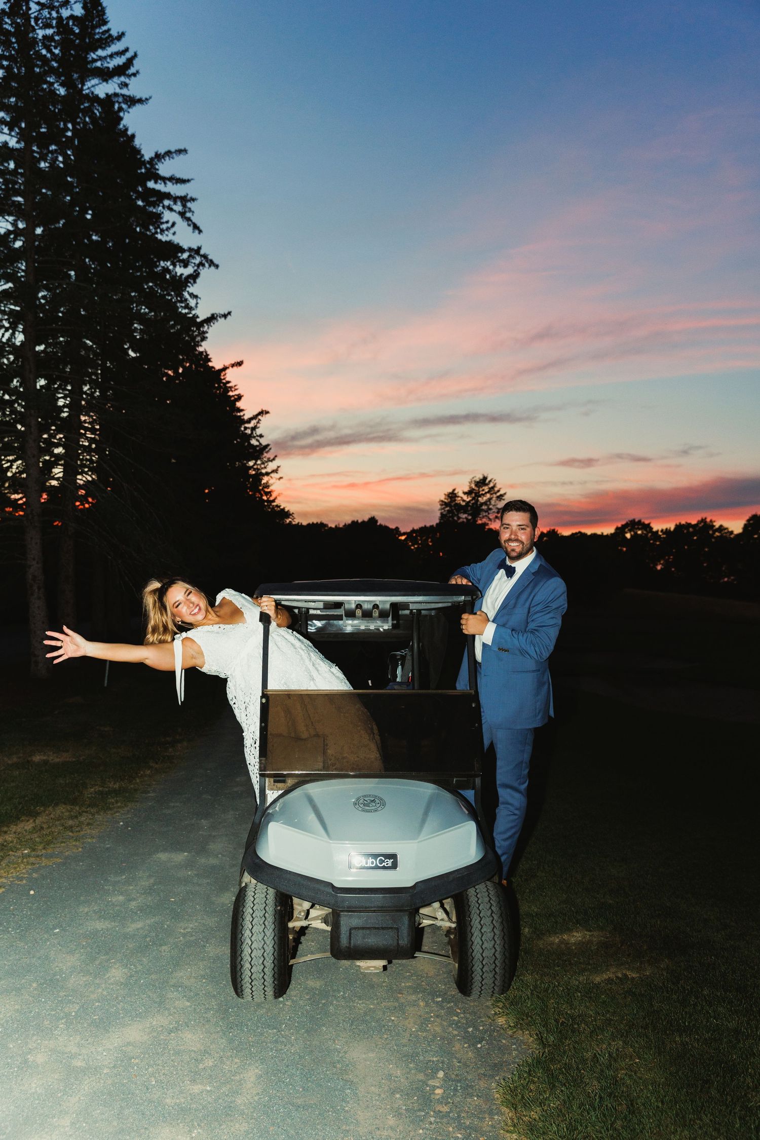 Wedding couple poses playfully with golf cart at sunset.