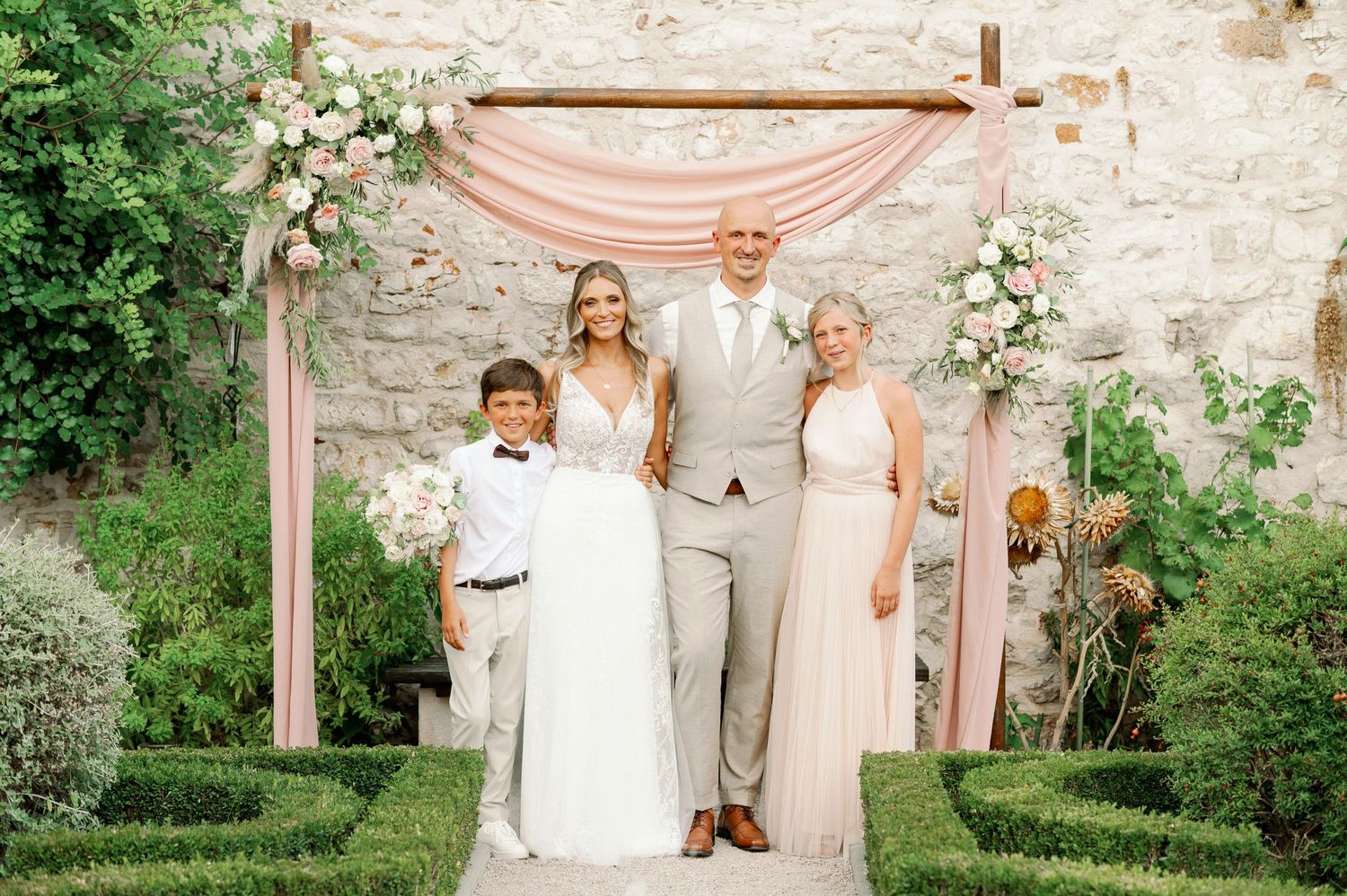 Wedding party poses under a blush pink archway against a stone wall backdrop.