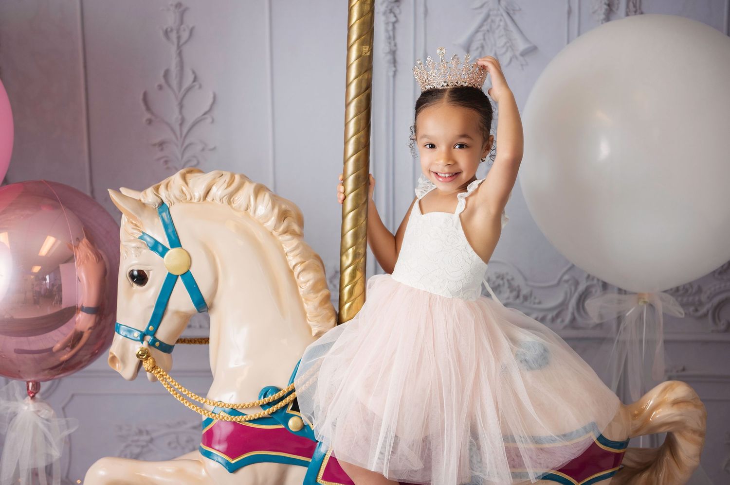 Adorable little girl in pink tutu and crown riding a decorative carousel horse with balloons.