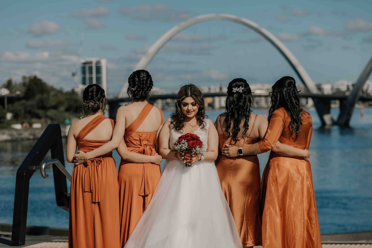 Bride in white dress with bridesmaids in orange dresses standing by waterfront with bridge view.
