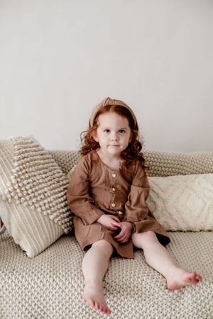 Child in brown dress sitting comfortably on textured beige couch with decorative pillows.