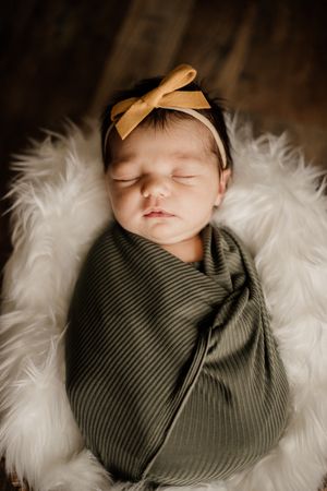 A sleeping newborn wrapped in olive green swaddle on white fur with a bow headband.
