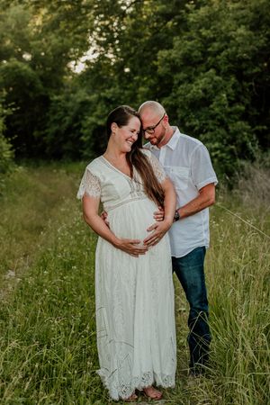 Maternity photos of couple in white dress and casual wear standing in tall grass field.