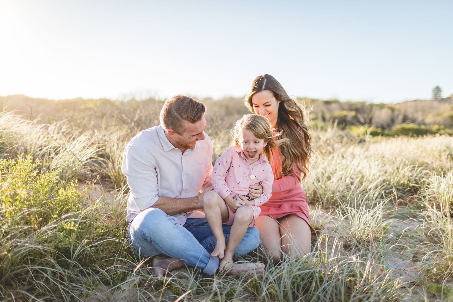 Family sitting together in golden sunlit field of tall grass during sunset photoshoot.