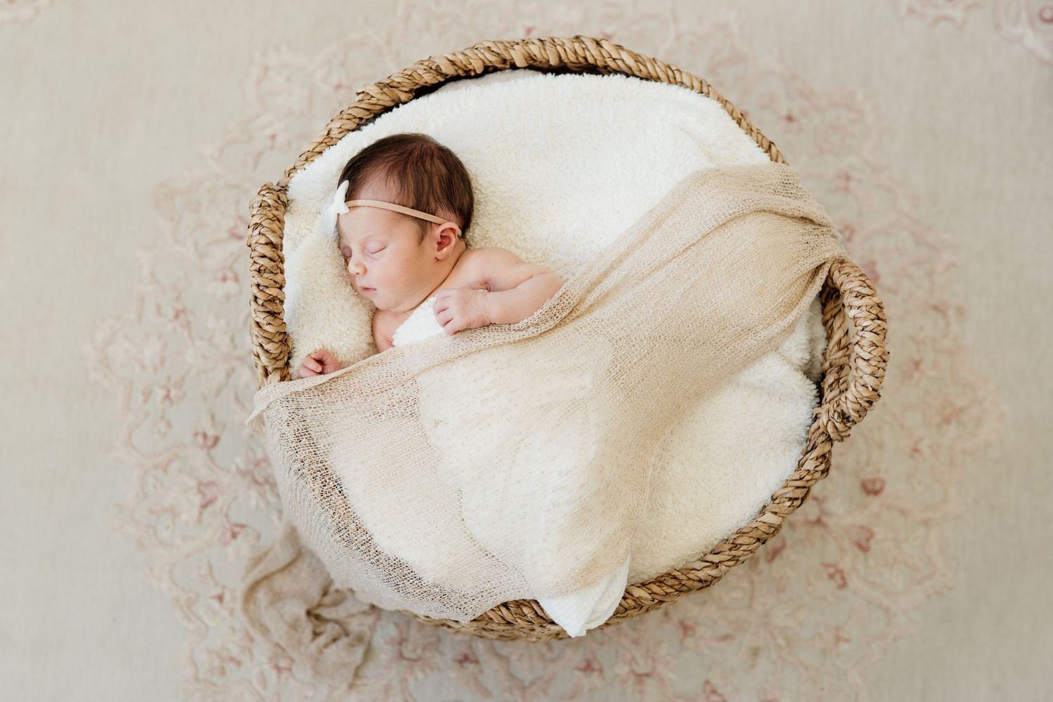 Newborn baby sleeping peacefully in round woven basket wrapped in soft white blanket against neutral background.