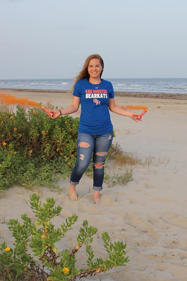 A person in a blue university t-shirt and distressed jeans poses on a beach with sand dunes and ocean in background.