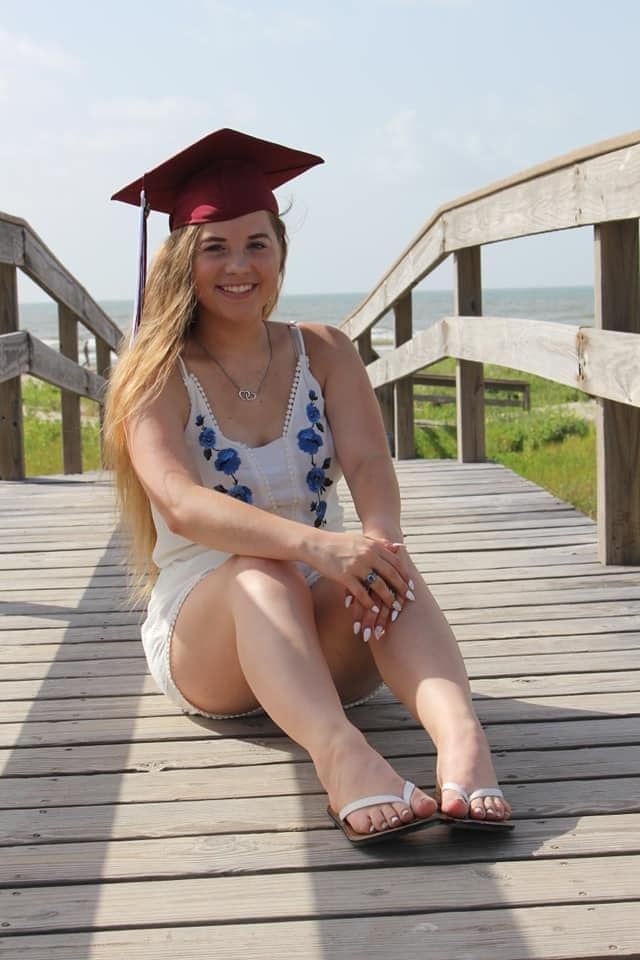 Graduate in red cap and white floral dress sitting on wooden boardwalk by the beach.