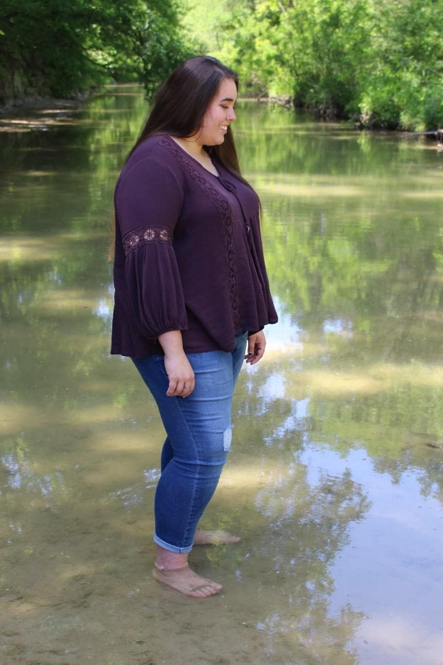 Person in purple bell sleeve top and jeans standing by tranquil pond reflection.