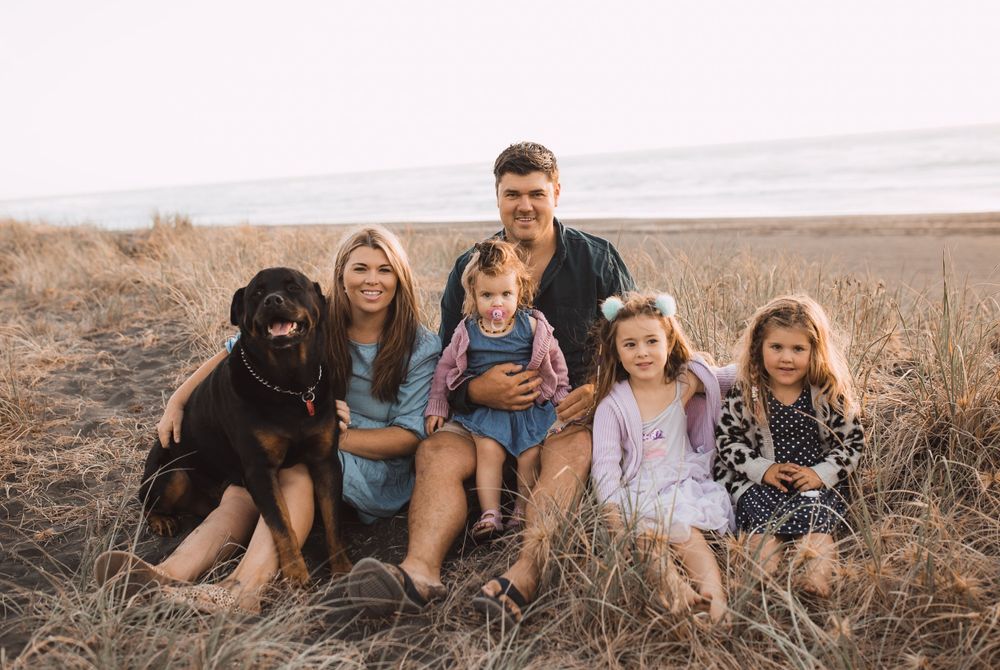 Family portrait with black lab dog sitting together in coastal grass at sunset.