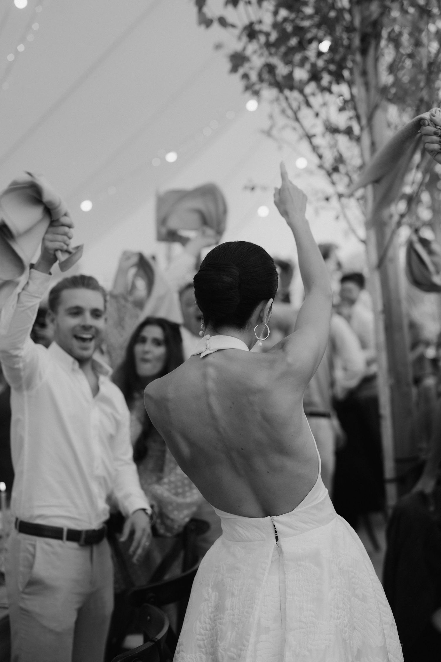 back of brides dress as she enters the marquee with guest cheering and waving napkins