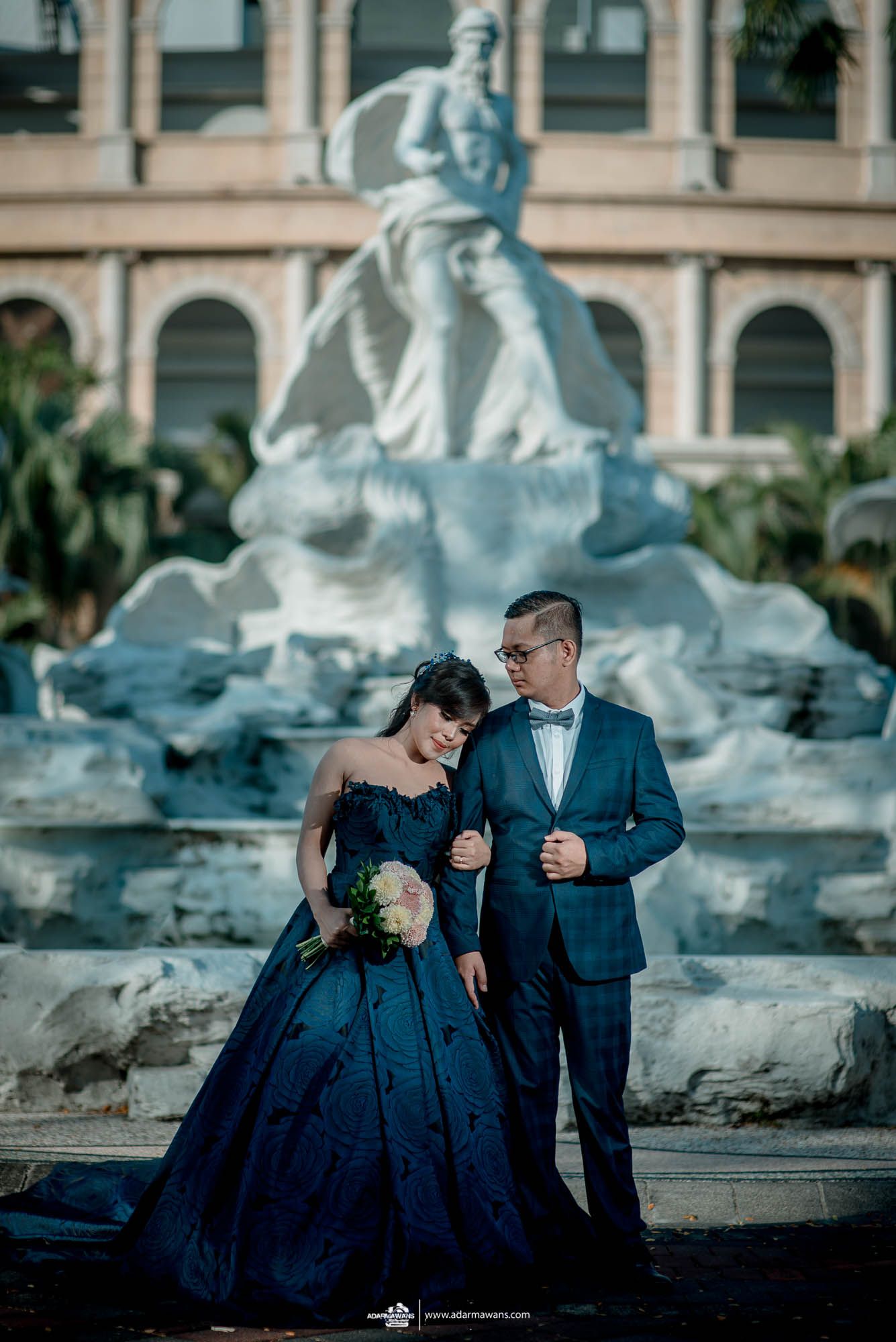 Elegant couple in formal attire posing by ornate fountain statue in classic setting.