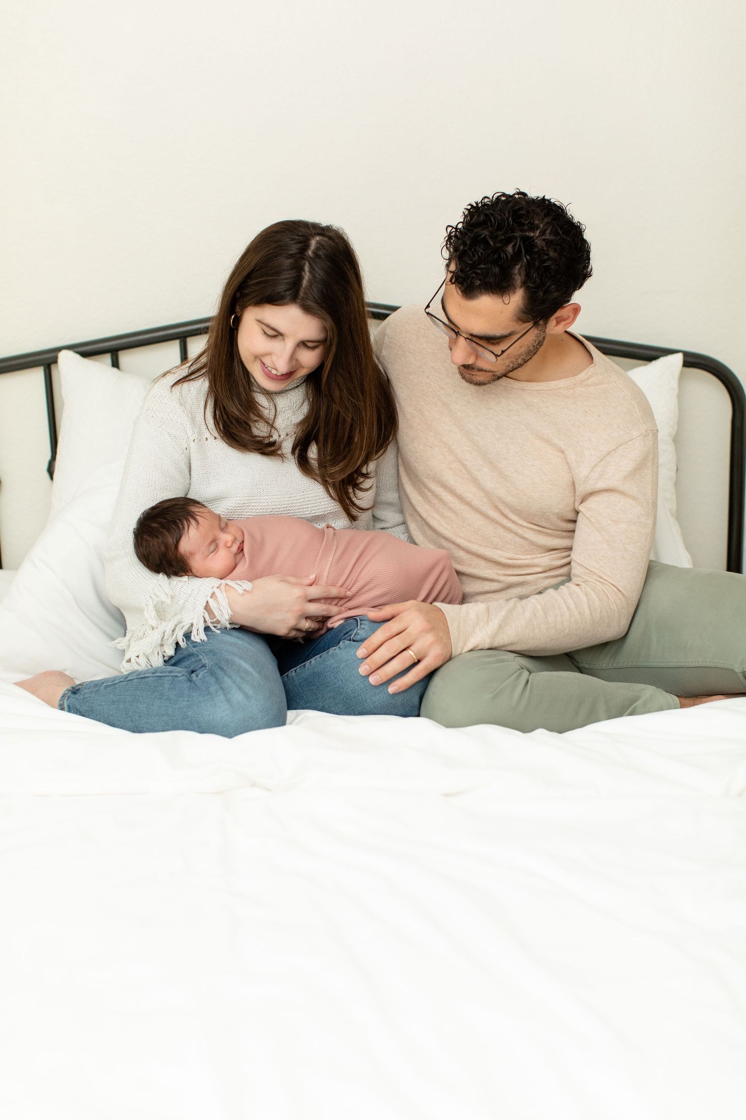 Parents cuddle with their newborn baby on a white bed with black metal headboard.