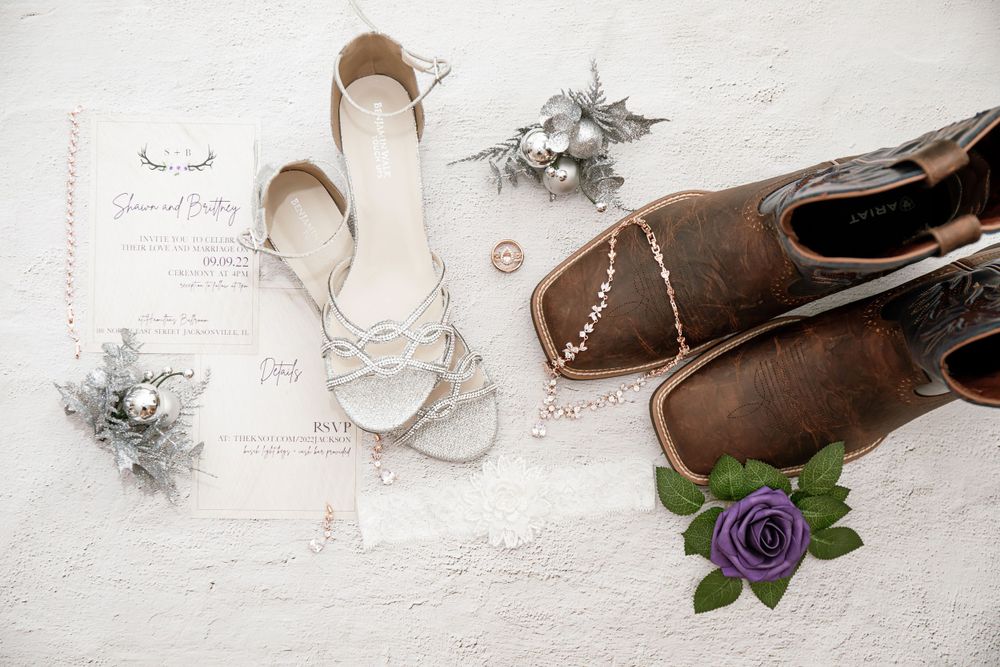 Wedding accessories including silver shoes, brown boots, and purple flowers arranged with invitation on white surface.
