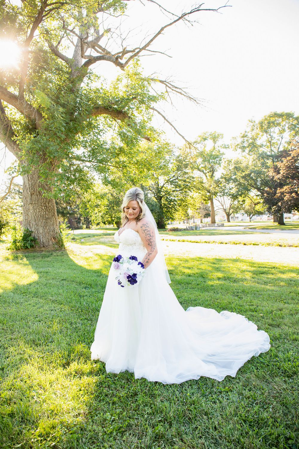A bridal portrait showing a flowing white gown and purple bouquet in a sunlit garden setting.