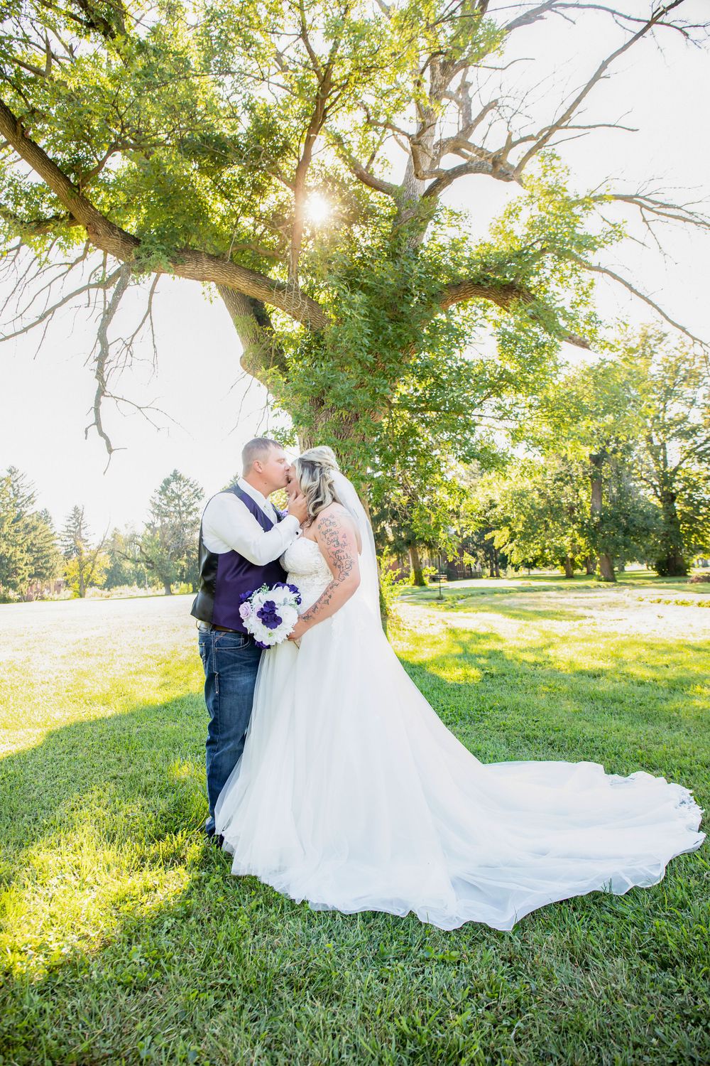 A romantic wedding portrait under a sunlit tree with purple bouquet details.