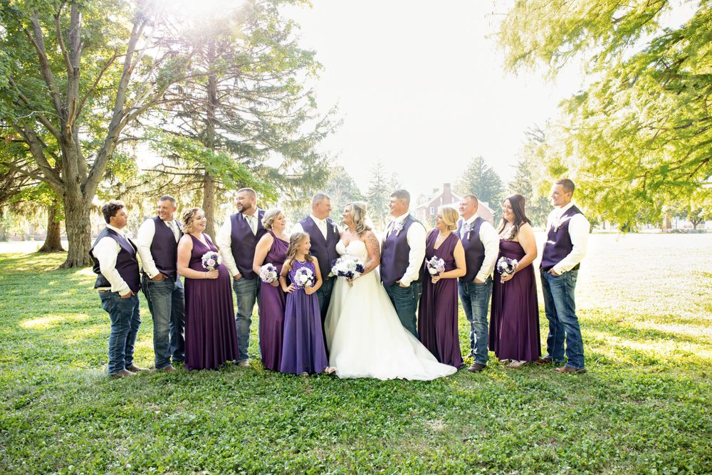 Wedding party in purple and navy attire poses together on a sunny lawn with trees in background.