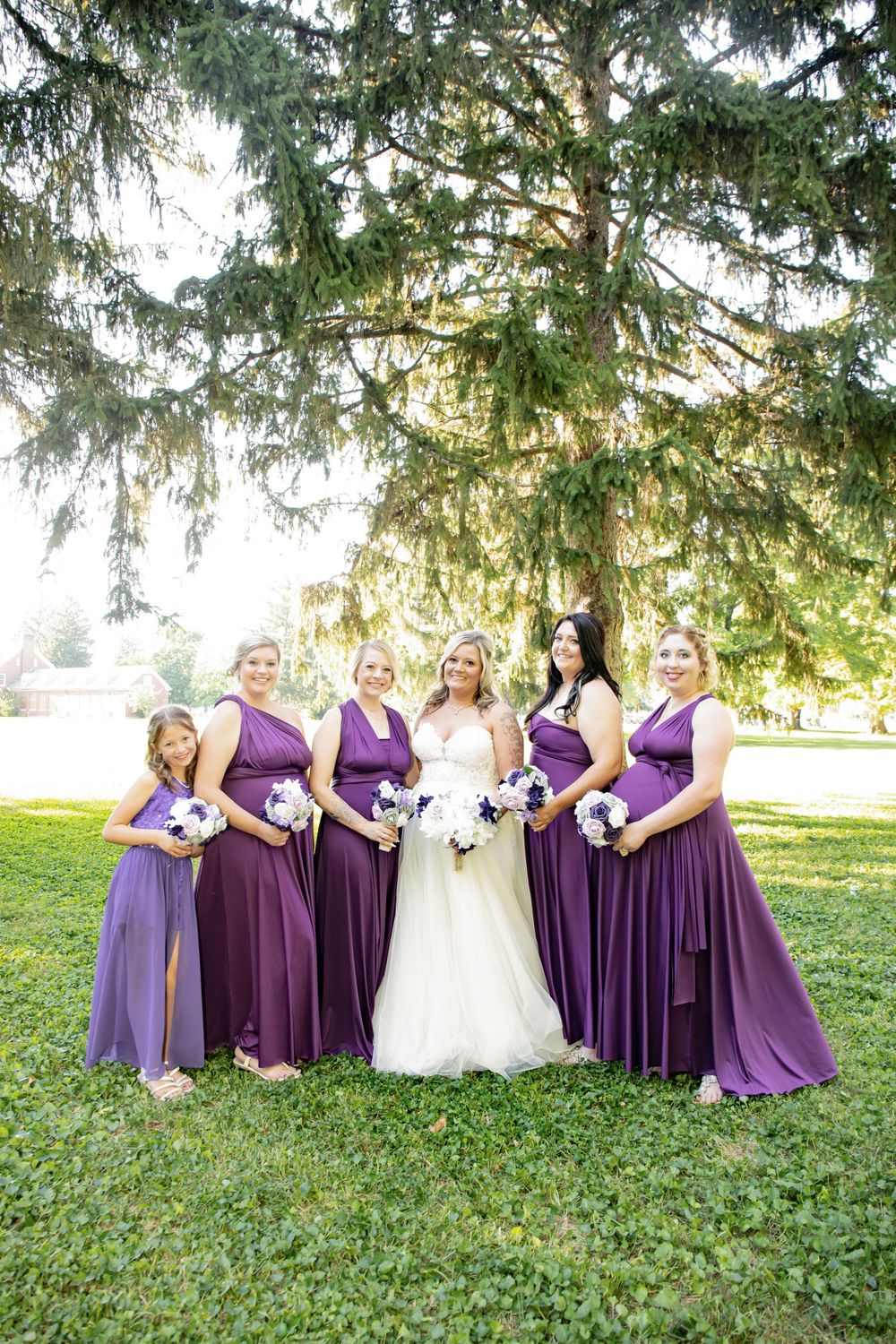 Bridal party in purple bridesmaid dresses poses together under a large tree on a sunny day.