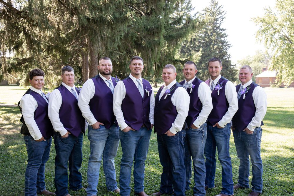 Groomsmen in matching purple vests and jeans pose together outdoors under evergreen trees.