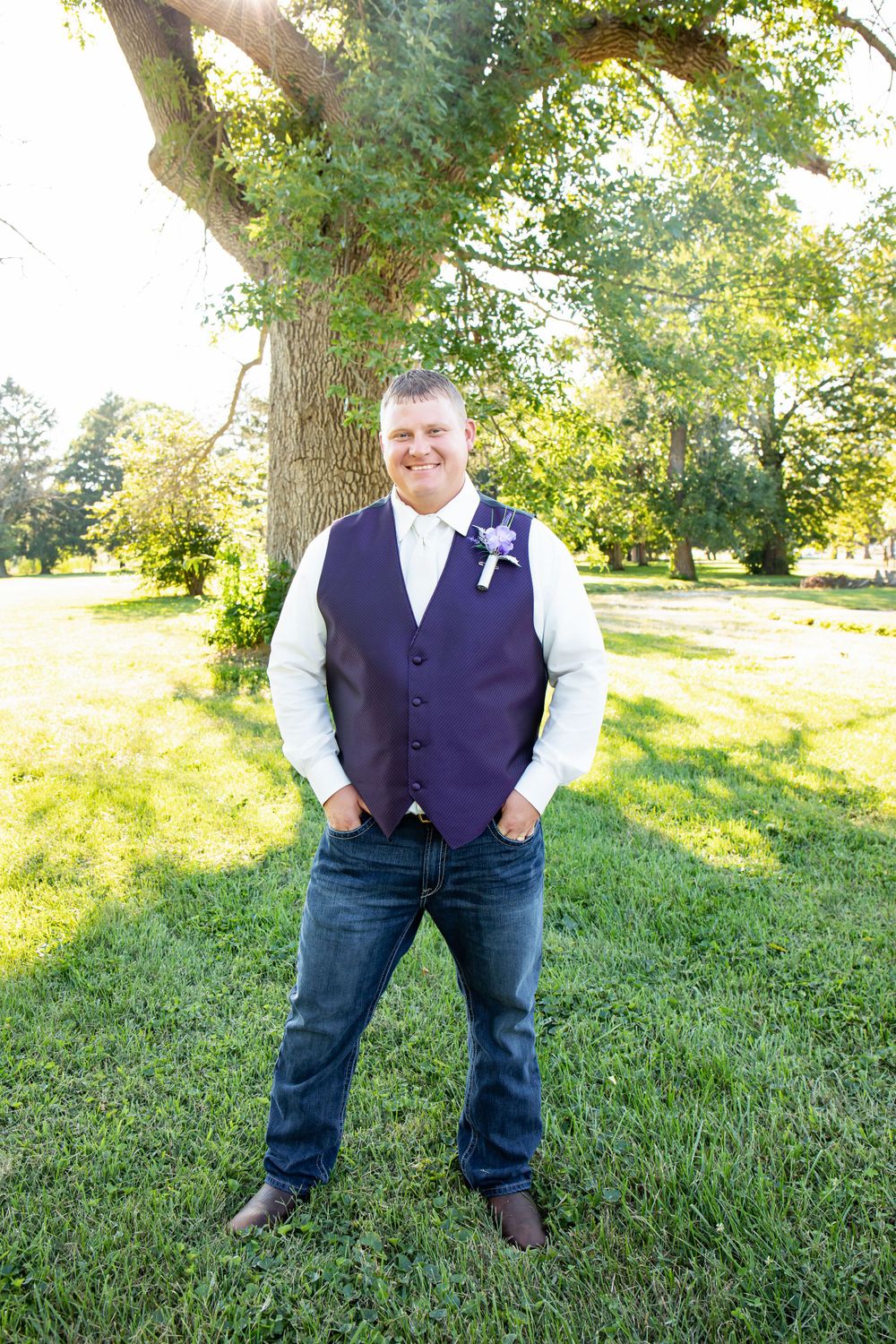 Person in navy vest and white shirt stands on grass under sunlit tree.