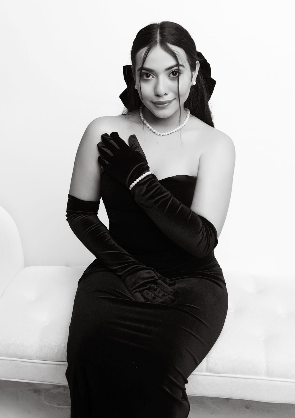 Black and white photo of an elegant woman in velvet dress seated on white furniture.