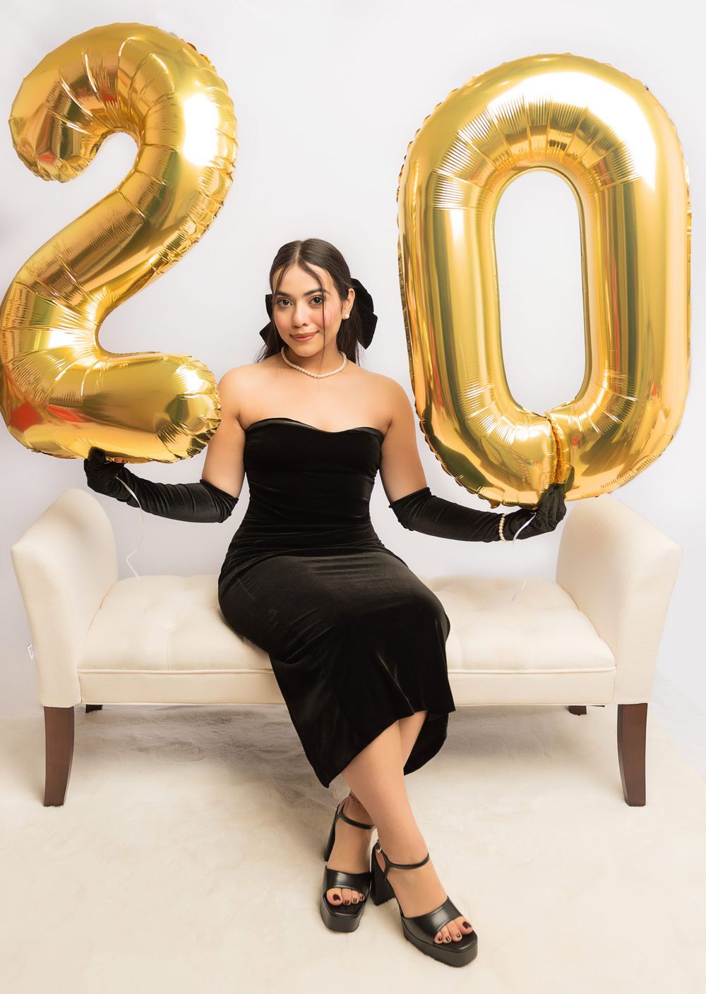 Woman in black dress poses with large gold number 20 balloons on white bench.