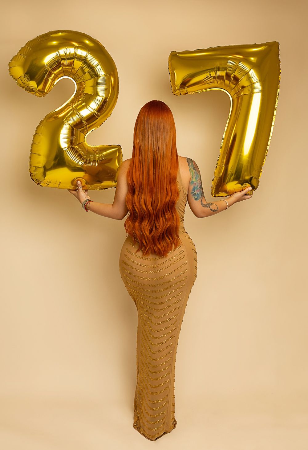 Person with long red hair holds golden number balloons 2 and 7 while wearing a fitted tan dress.