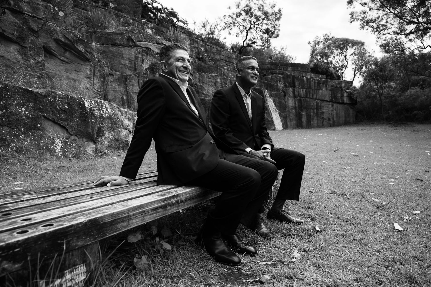 Two men in suits sitting casually on wooden bench near stone wall.