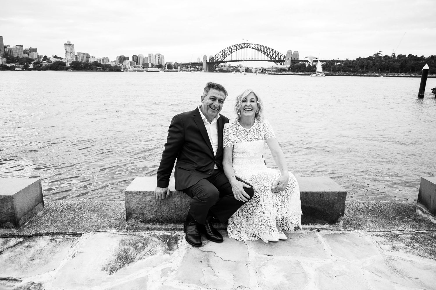 A black and white photo of a couple sitting by Sydney Harbour with the iconic bridge in the background.