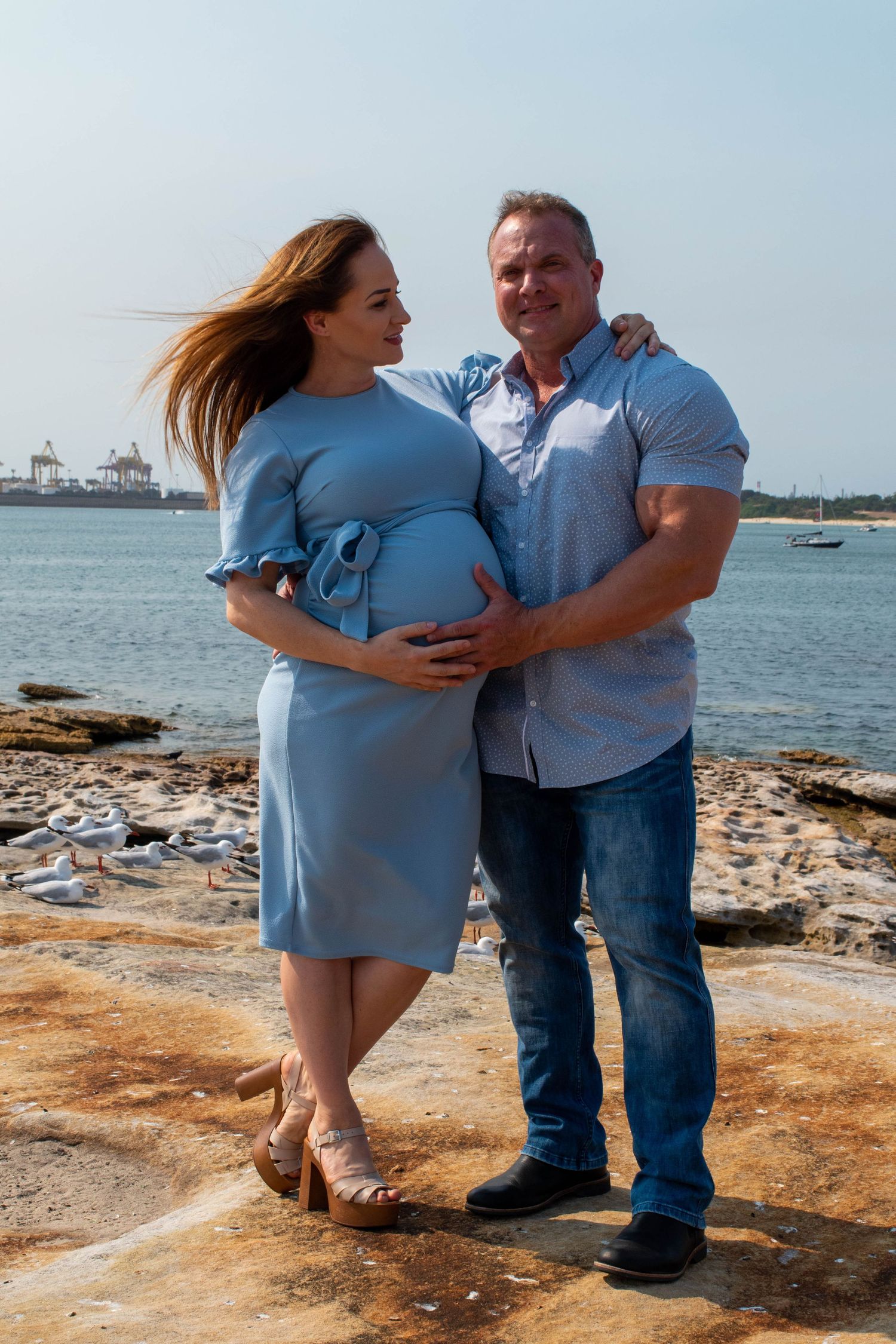 A maternity photo of a couple posing by the waterfront on a sunny day.