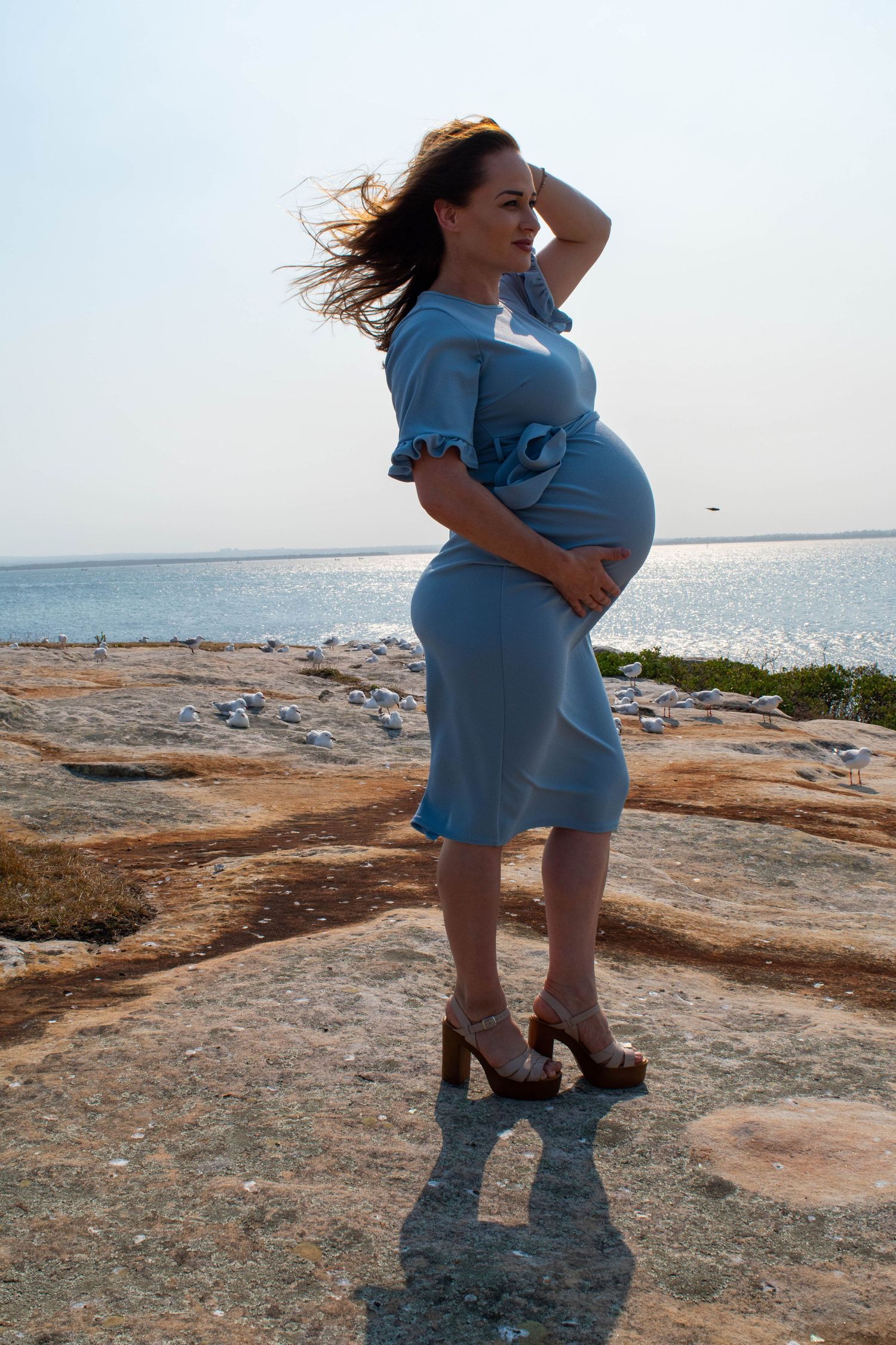 Woman in a blue dress posing on rocky coastline during maternity photoshoot.