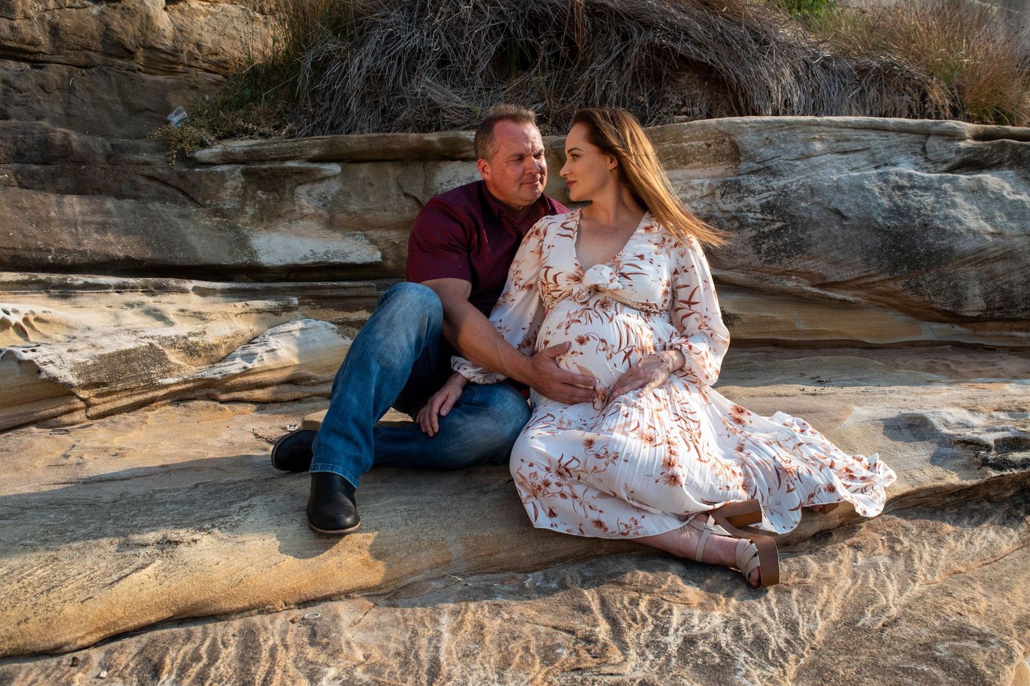 Romantic maternity photoshoot on rocky cliffs showcases couple sitting together on natural stone formations.