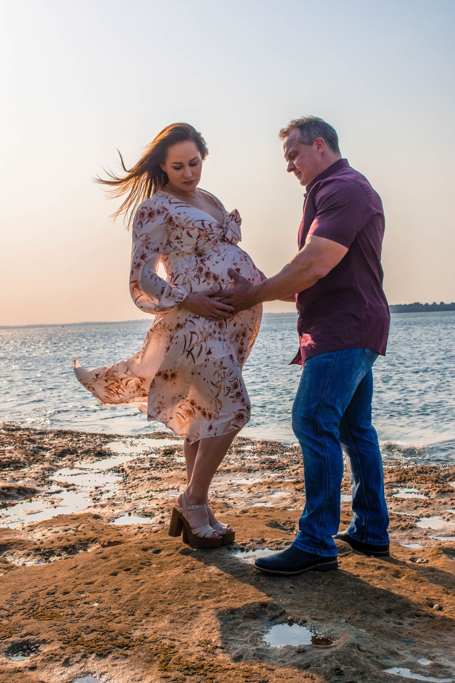 Romantic beach maternity photo with flowing floral dress catching the wind at sunset.