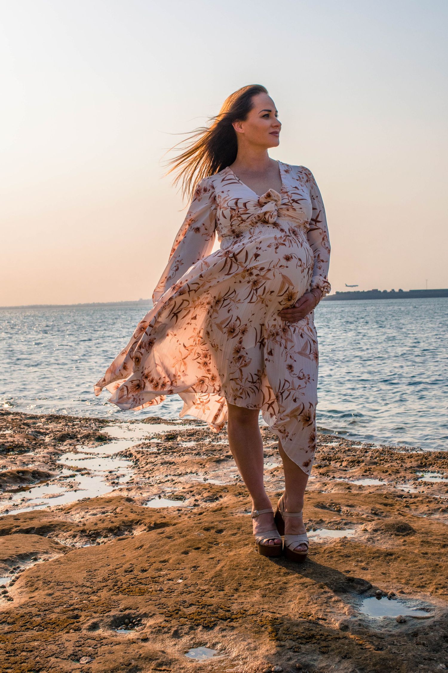Pregnant woman in floral dress stands on beach during golden hour as wind catches her dress.