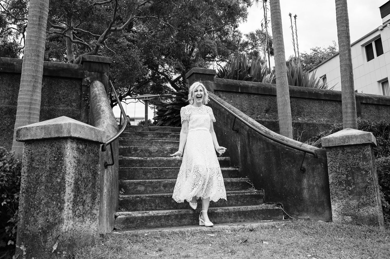 Woman in white dress poses joyfully on stone steps between palm trees.