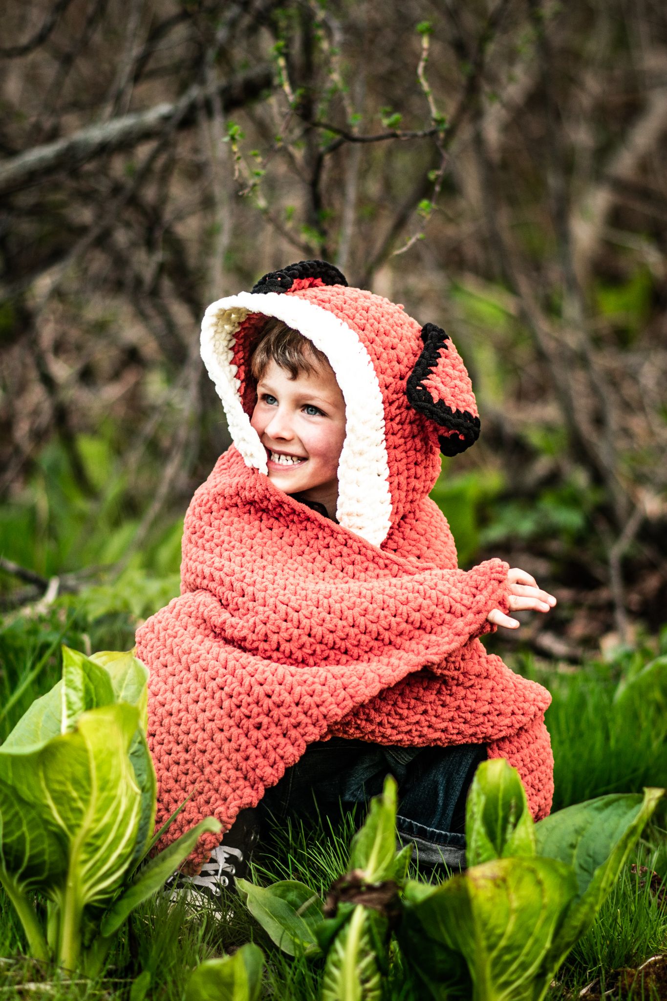 A child wrapped in a cute polka dot fox hooded blanket sits among green plants.