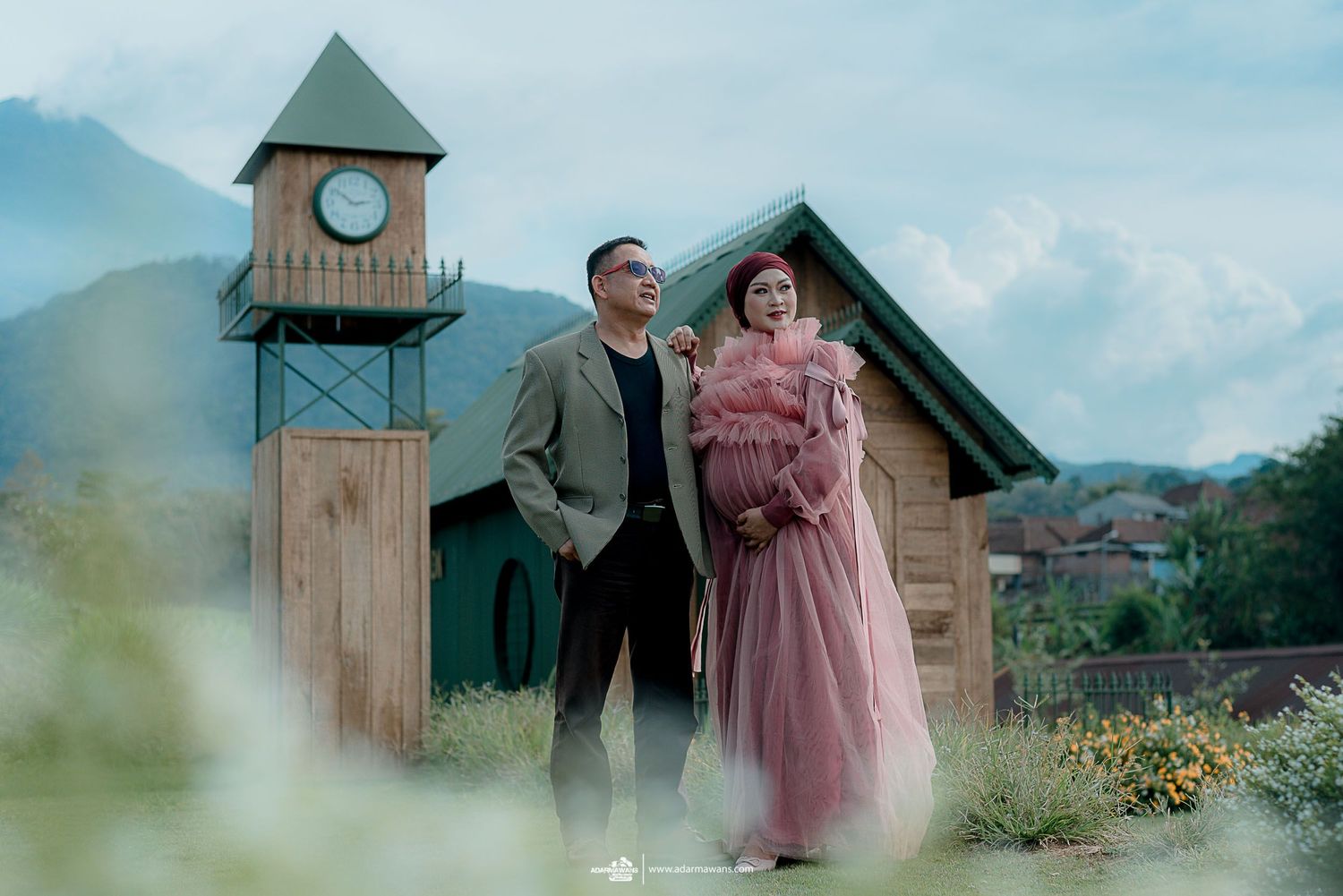 Couple in vintage-inspired outfits posing near a rustic clock tower and mountain backdrop.